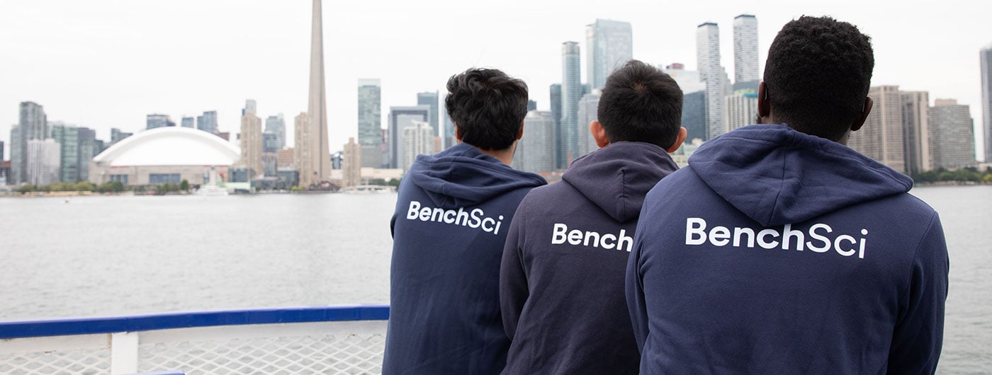 Three people wearing navy blue "BenchSci" hoodies stand at the railing of a boat, looking out at the Toronto skyline.