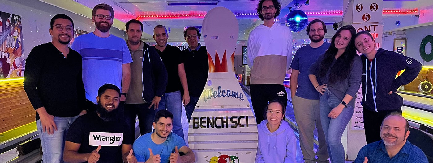 BenchSci team members pose for a photo at a bowling alley. They are gathered around a large decorative bowling pin that says "Welcome BenchSci" under glowing neon lights.